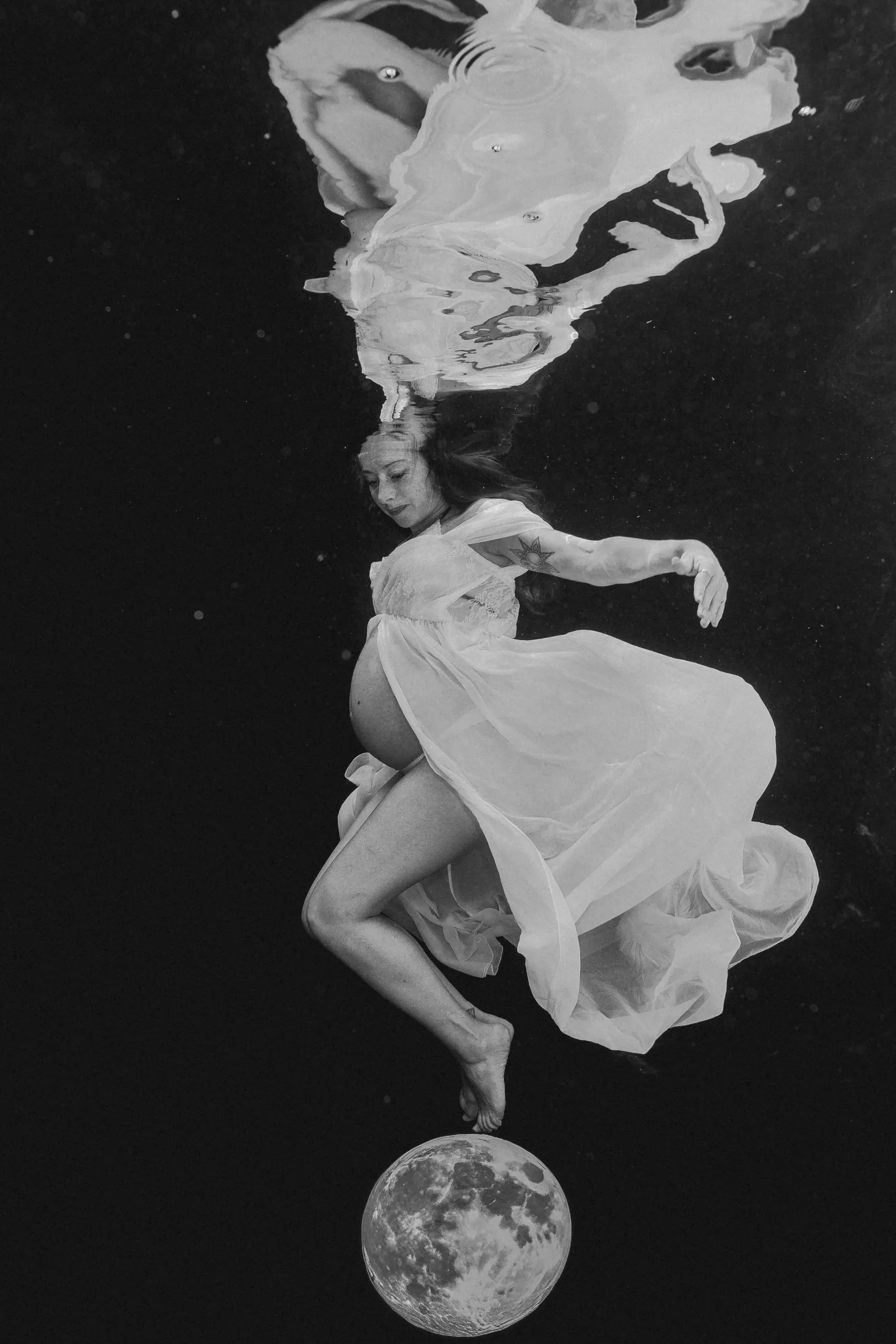 Black and white underwater portrait of a pregnant woman gracefully dancing on the moon during her babymoon.