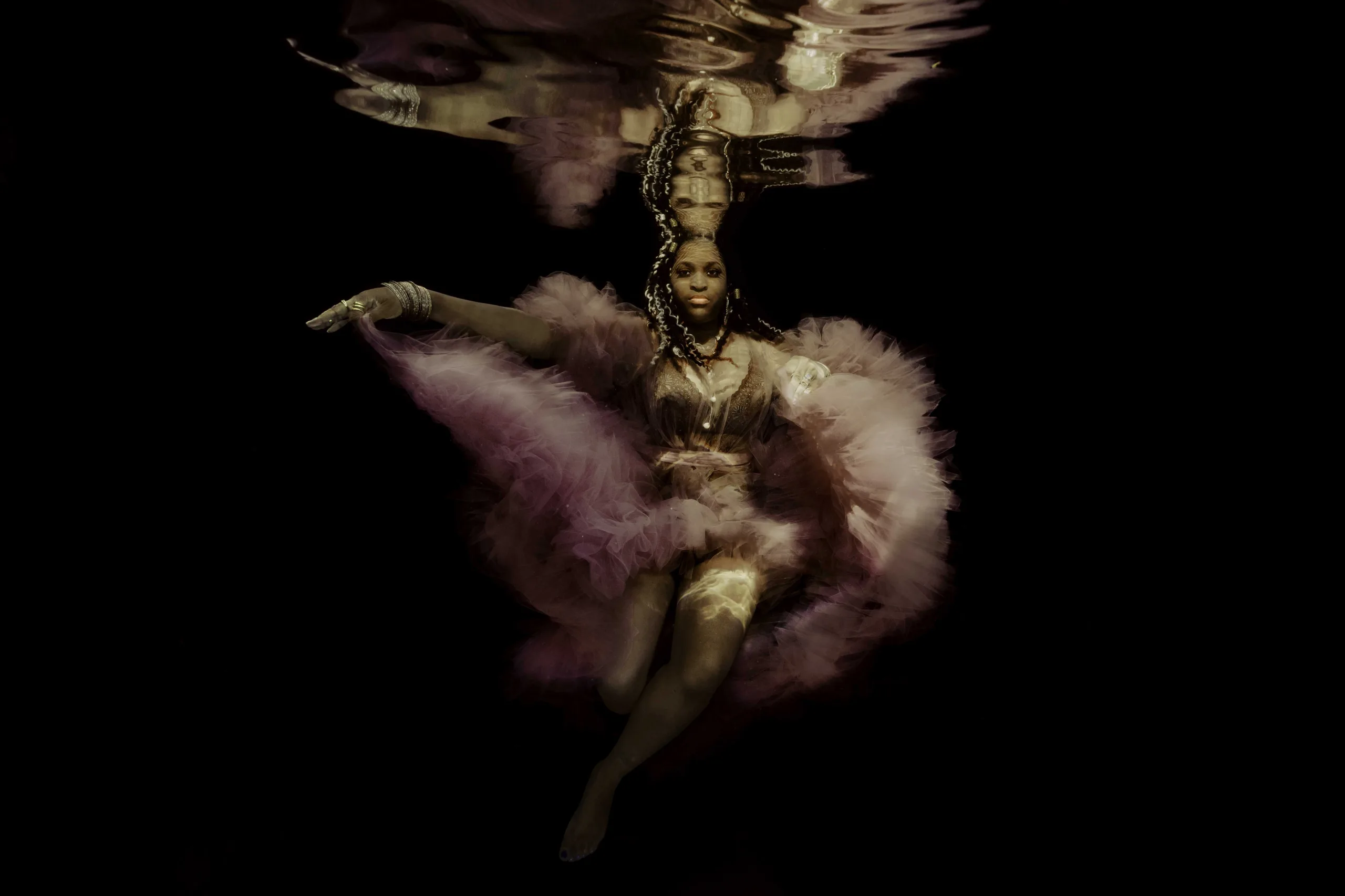 Afro-American woman gracefully posing underwater, wearing a flowing pink ballet dress with soft movements, surrounded by water.