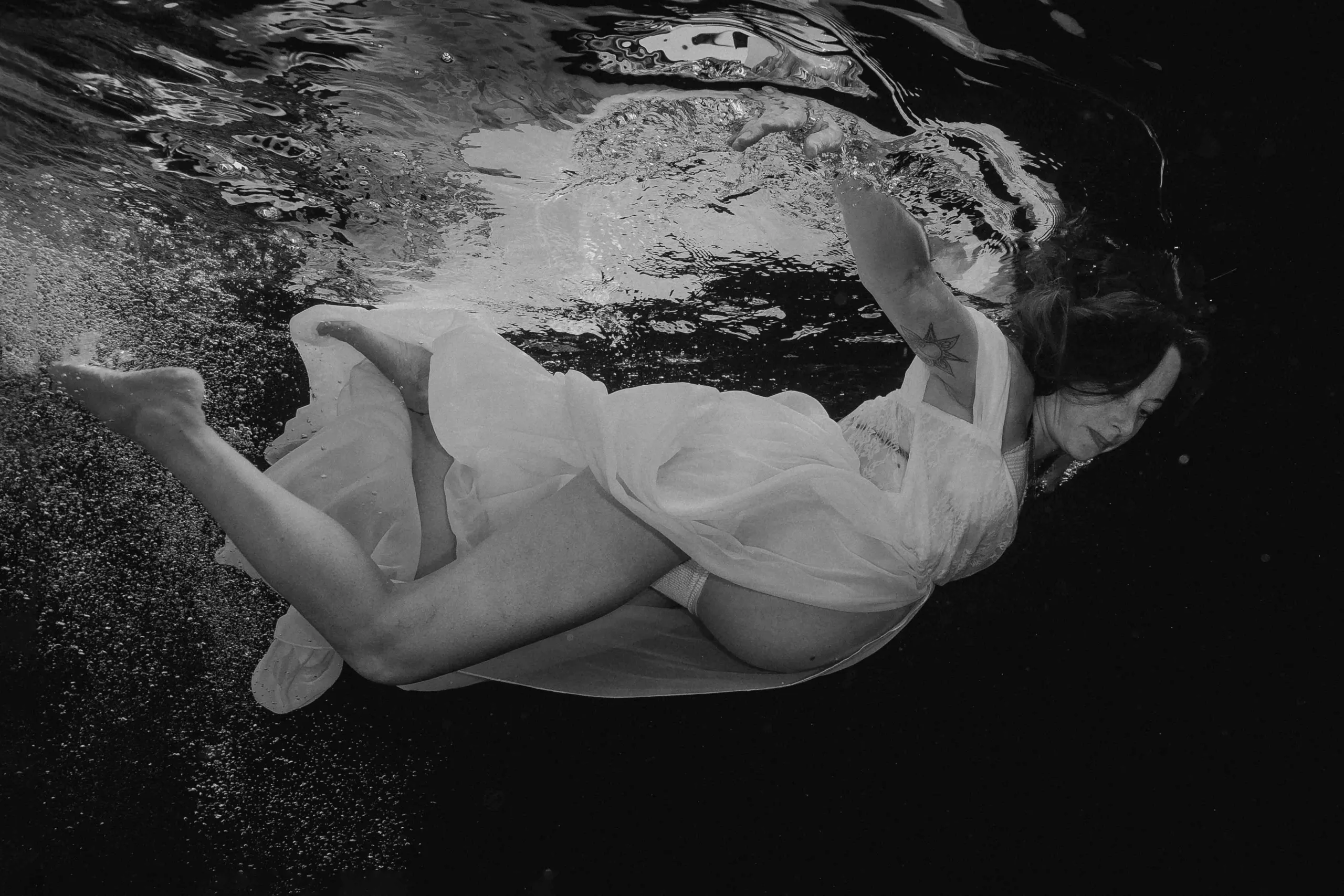 A pregnant woman swims underwater in a flowing white dress. The fabric billows around her, creating a sense of fluidity and grace. Her serene expression and calm posture reflect peace and anticipation. Light filters through the water, creating a dreamy, ethereal atmosphere.