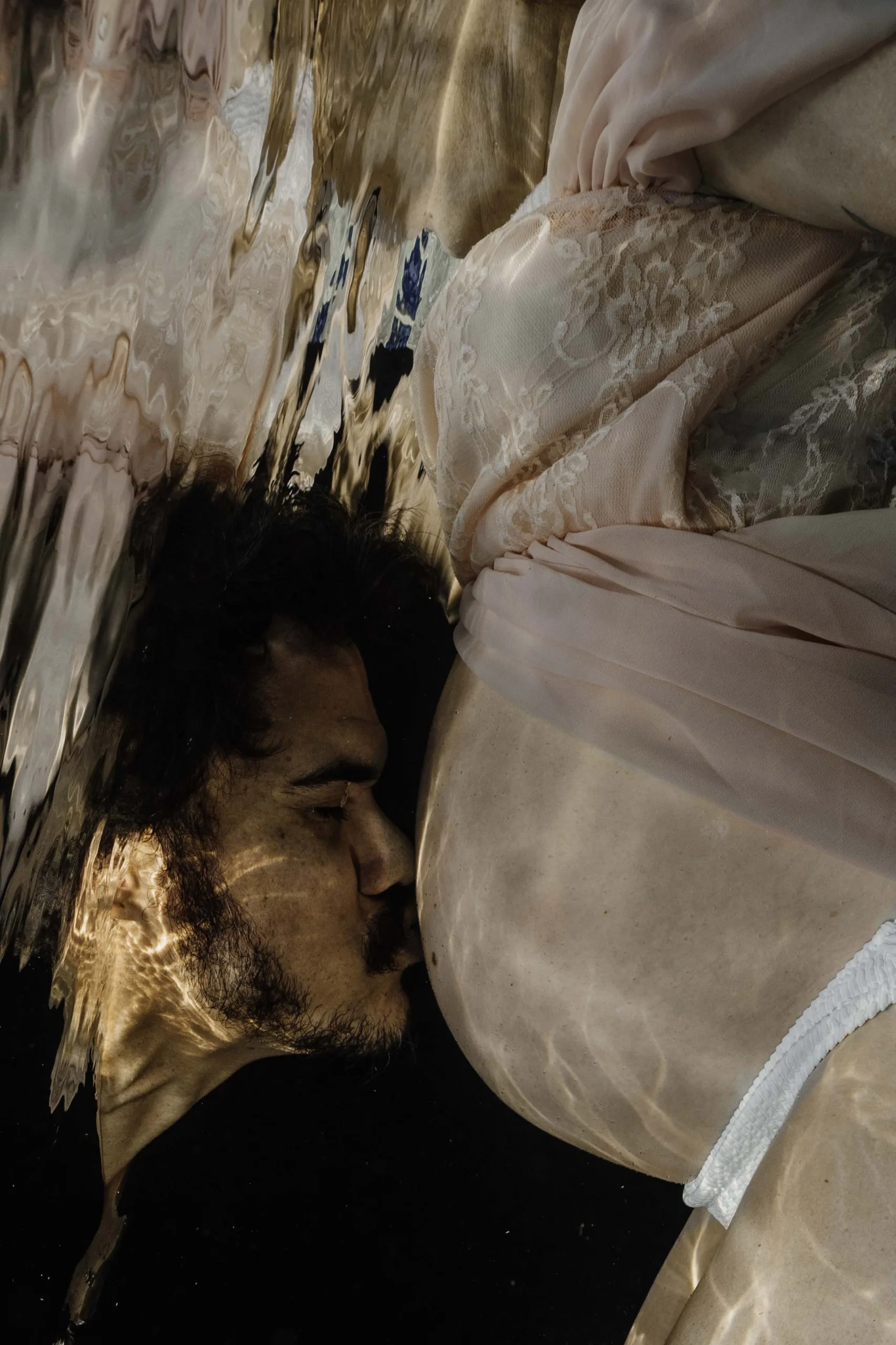 A man gently kisses the pregnant belly of his partner underwater.