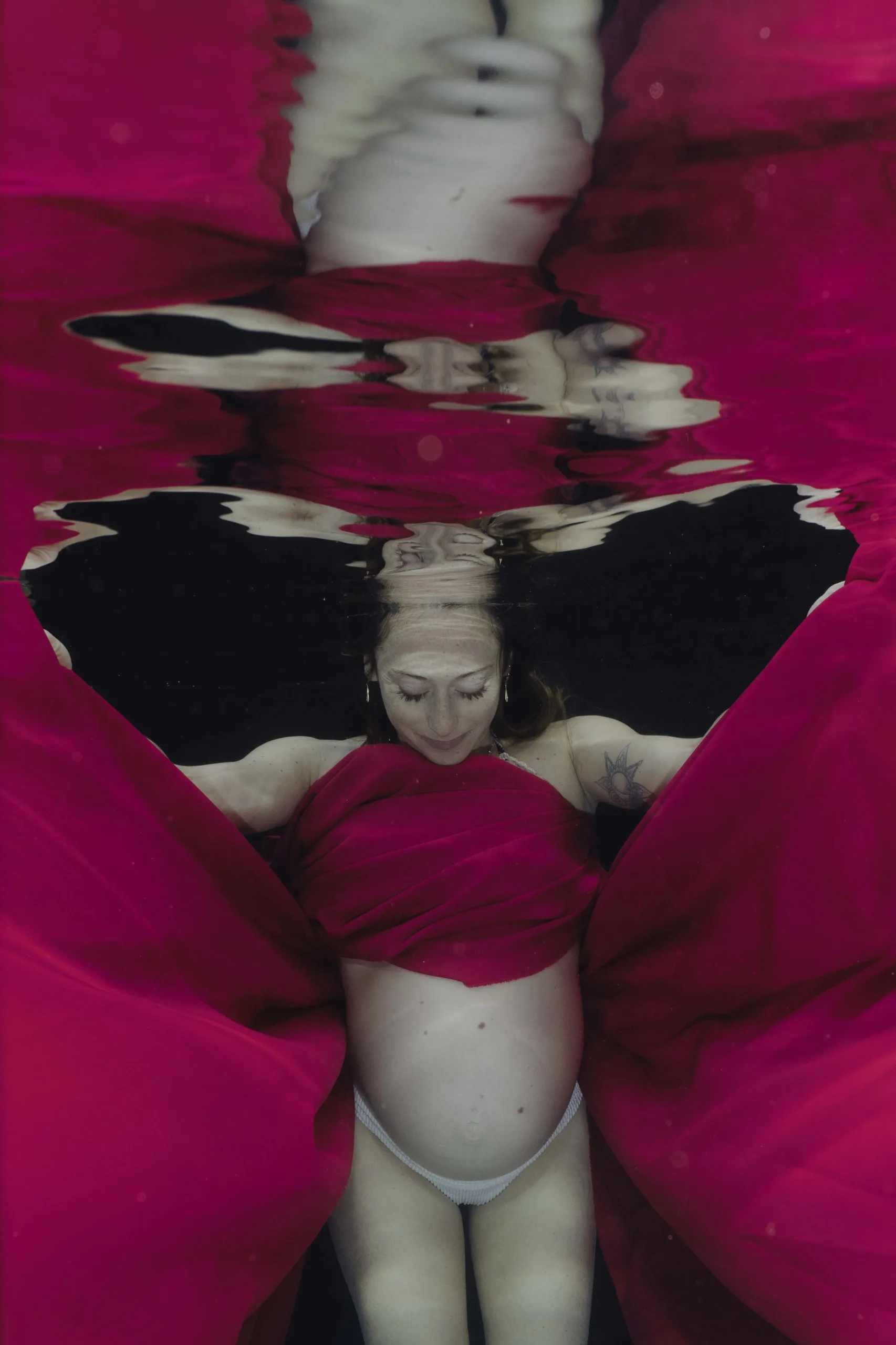 A pregnant woman underwater, wrapped in pink fabric that moves around her gracefully.