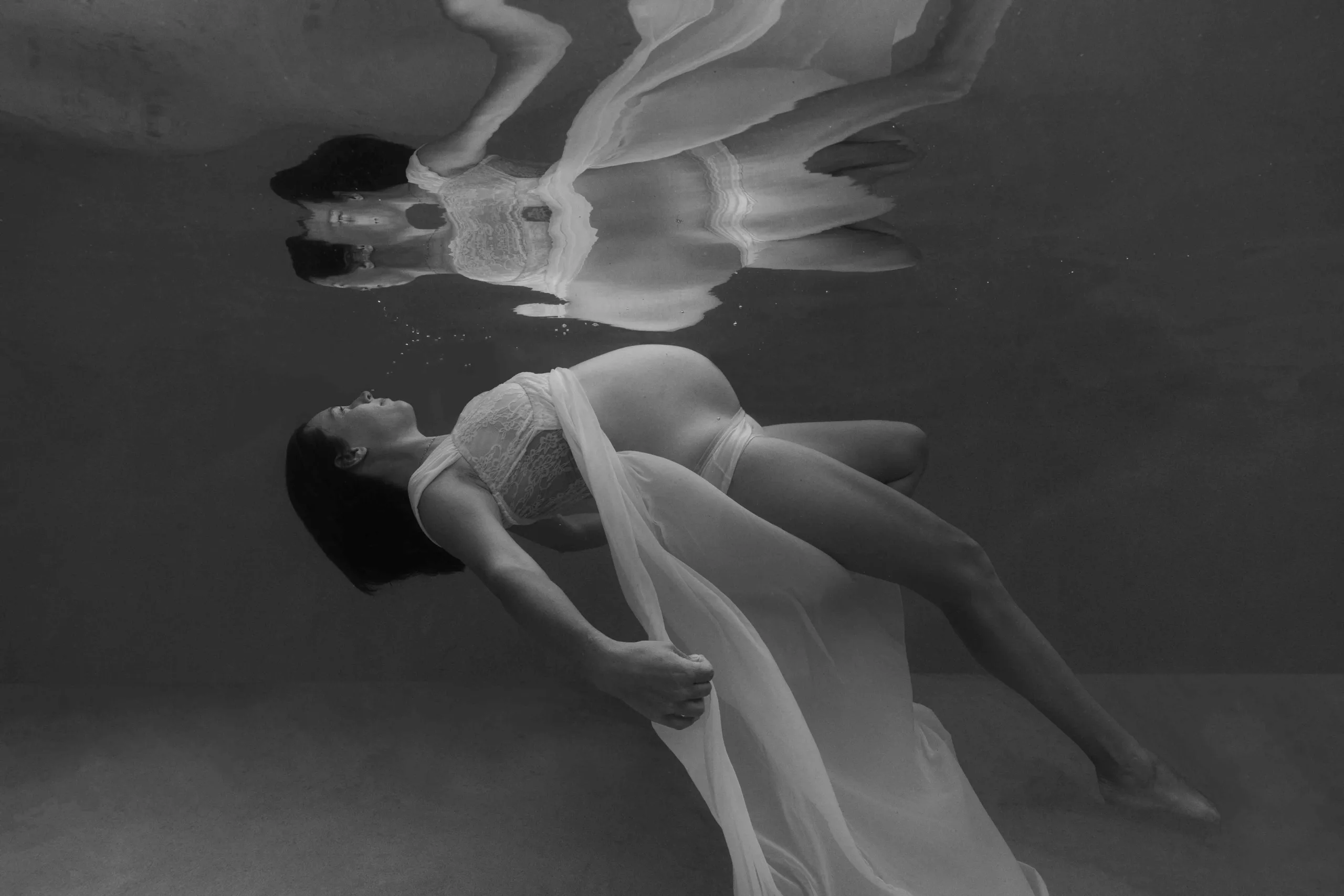 A pregnant woman floats gracefully underwater, wearing a flowing, sheer gown that moves around her in soft waves.